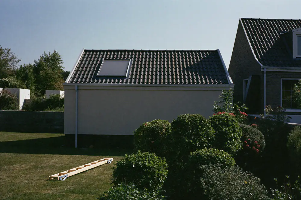 Achterdakvlak van een woning met donkere pannen en een dakraam, een ladder in het gras en de buurwoning ernaast