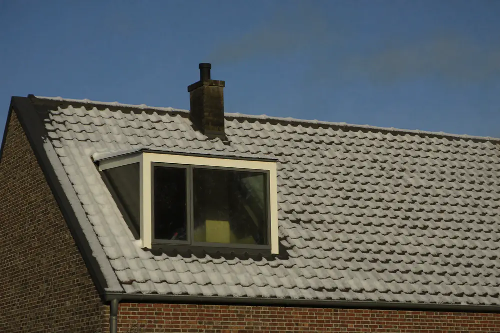 Pannendak met een laagje rijp en sneeuw, een dakkapel met grijze kozijnen en een schoorsteen met rook