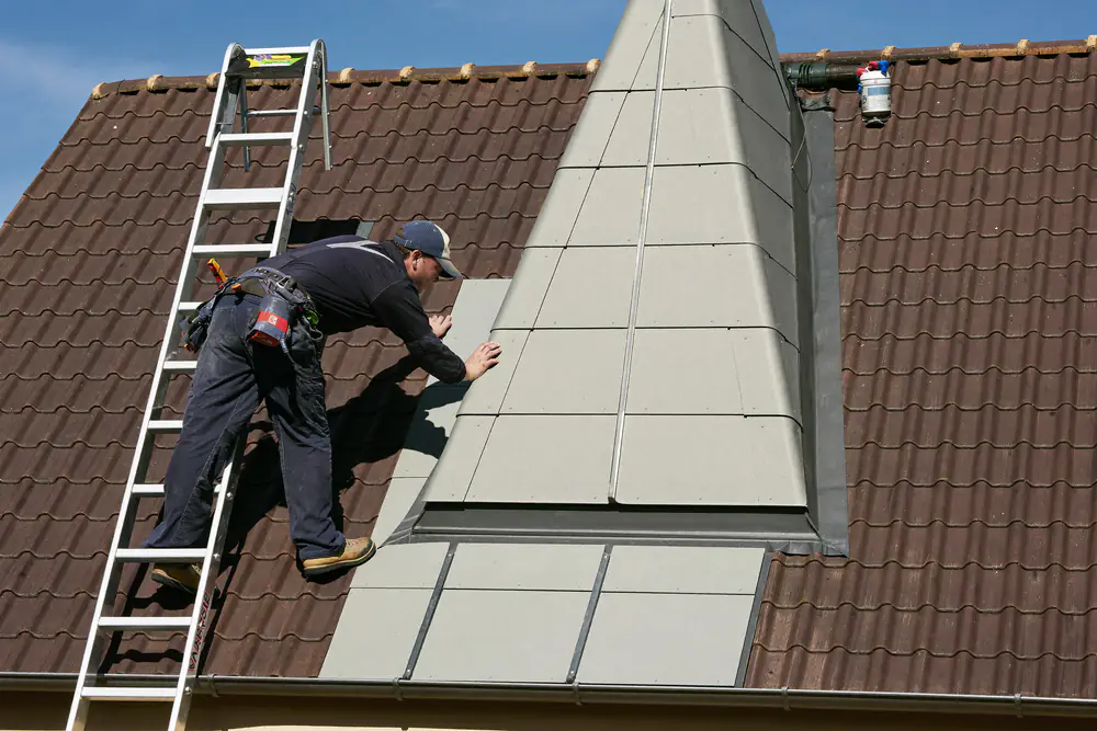 Dakdekker brengt grijze bekleding aan op de zijwang van een dakkapel, met een ladder tegen het dak