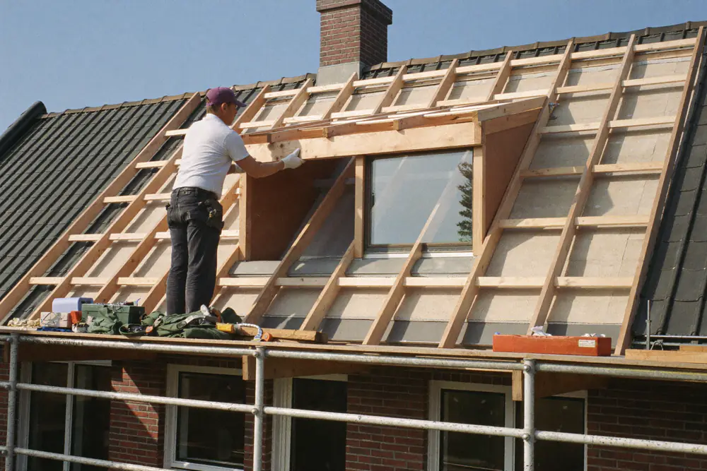 Timmerman bouwt een traditionele dakkapel op het dak, met zichtbare balken en isolatieplaten, naast een steiger