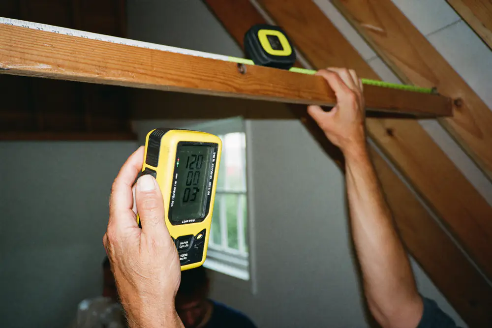 Handen met een laserafstandsmeter en een rolmaat tegen een houten dakspant in een zolder tijdens het opmeten