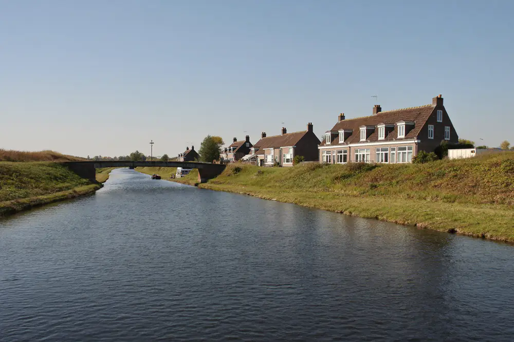 Recht kanaal in een vlak landschap met bakstenen woningen met dakkapellen langs de oever en een lage brug