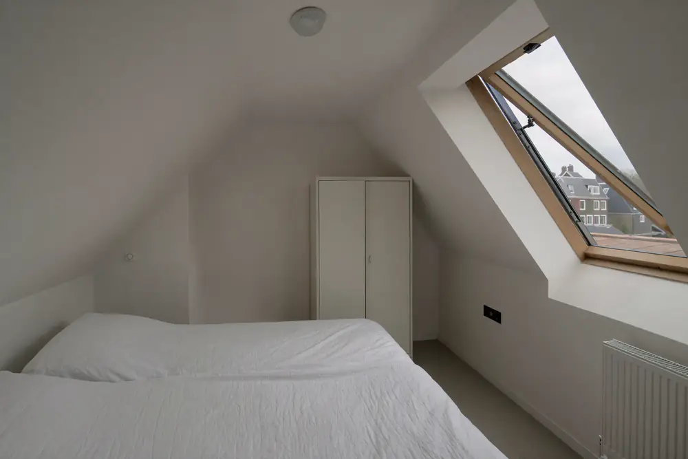 Verbouwde zolderkamer met een groot raam, een kast langs de knieschot en daglicht over de vloer