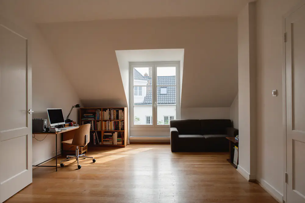 Zolderkamer met een dakkapelraam dat volle staanhoogte geeft, een bureau, een bank en een boekenkast